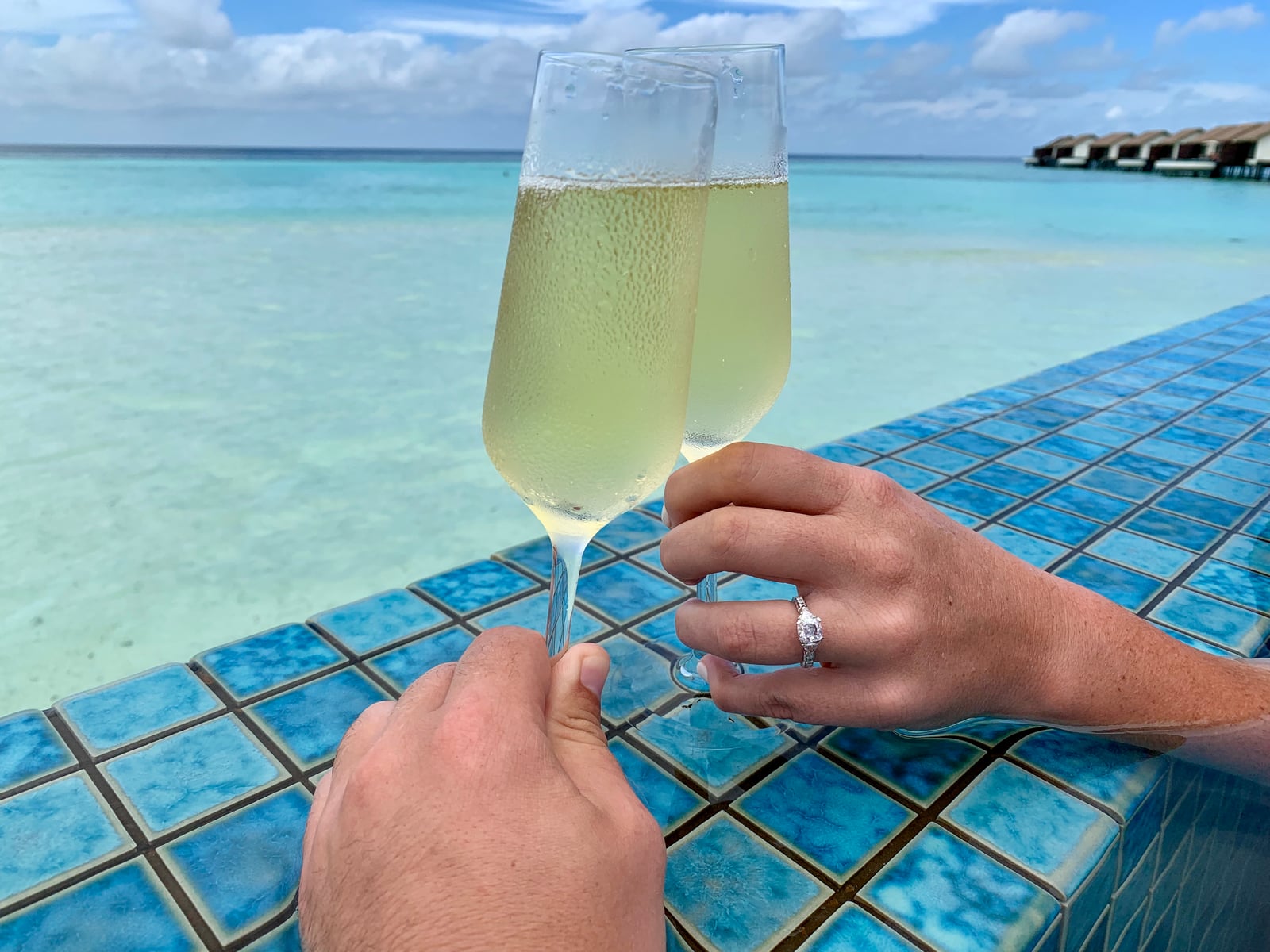 Two champagne glasses clinking on a pool deck with an engagement ring visible