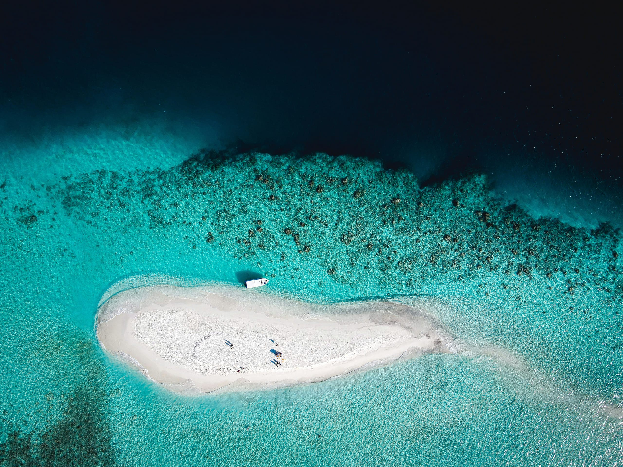 A boat moored at a bare white sandbank with a handful of people on it, turquoise lagoon all around, seen from above
