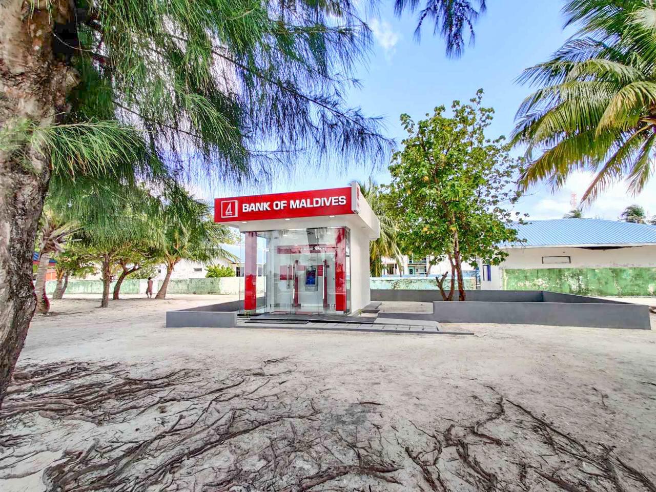 Un distributeur automatique de la Bank of Maldives sous des filaos et des palmiers, dans une rue sablonneuse d'une île locale