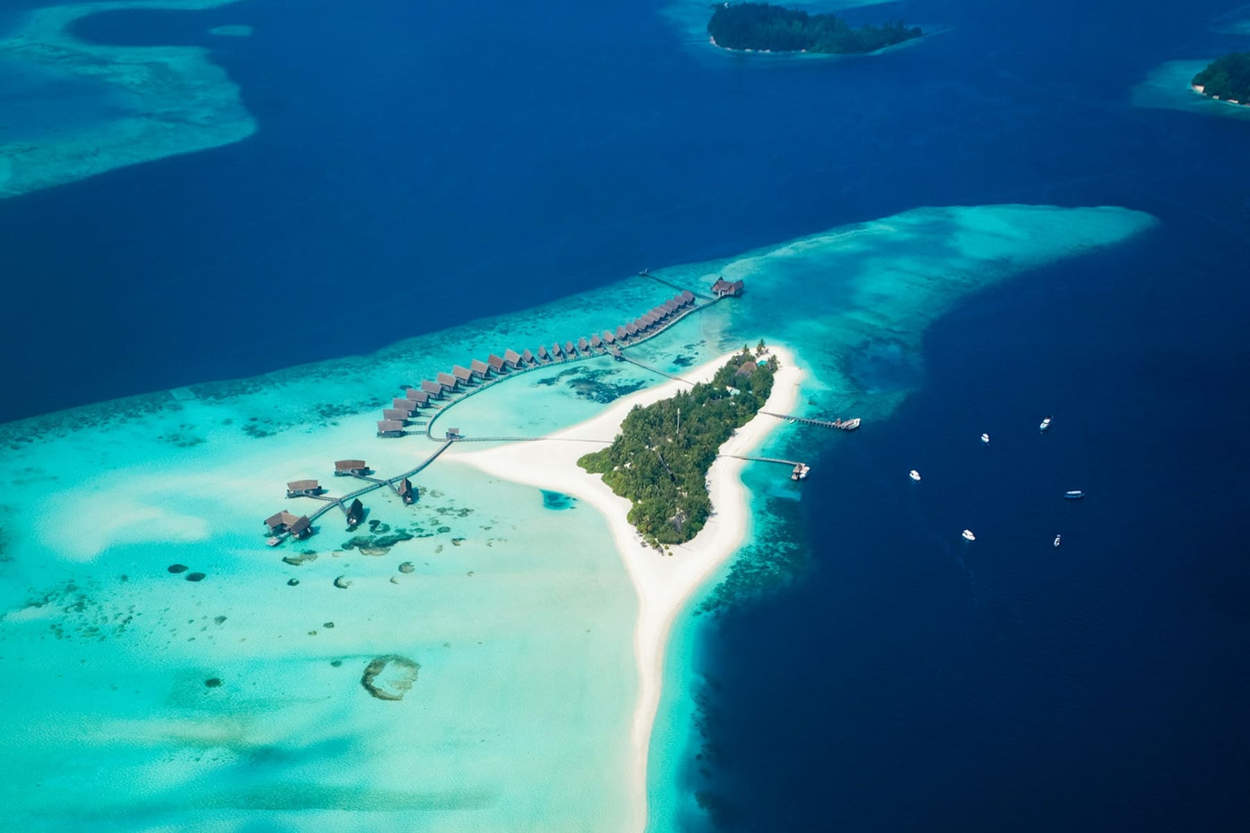 Vue aérienne de COMO Cocoa Island dans l'atoll de South Malé — une ligne courbe de villas sur pilotis, une petite île verte et un long banc de sable blanc dans un lagon turquoise cerné de chenaux bleu profond