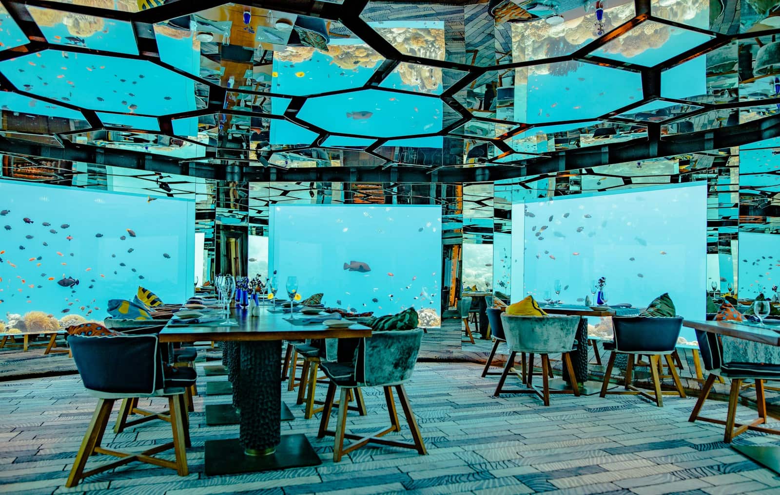 The interior of a Maldives undersea restaurant — set tables under a mirrored ceiling, reef and blue water visible through the glass walls