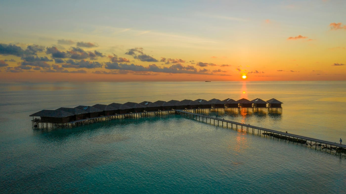 A row of overwater villas on stilts reaching into a calm sea at sunset, with a boardwalk