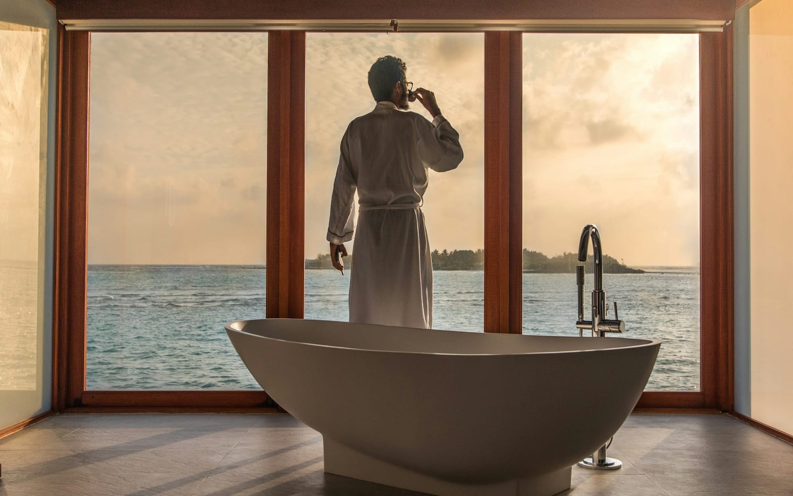 Un client en peignoir blanc devant les baies vitrées d'une villa sur pilotis aux Maldives, près d'une baignoire îlot autonome, contemplant le lagon au coucher du soleil