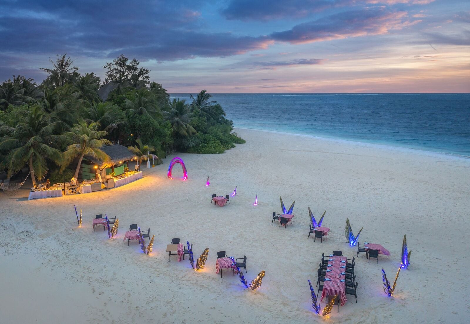Tische am Strand mit violetter Beleuchtung und einem dekorativen Bogen bei Sonnenuntergang in der Nähe des Resorts.