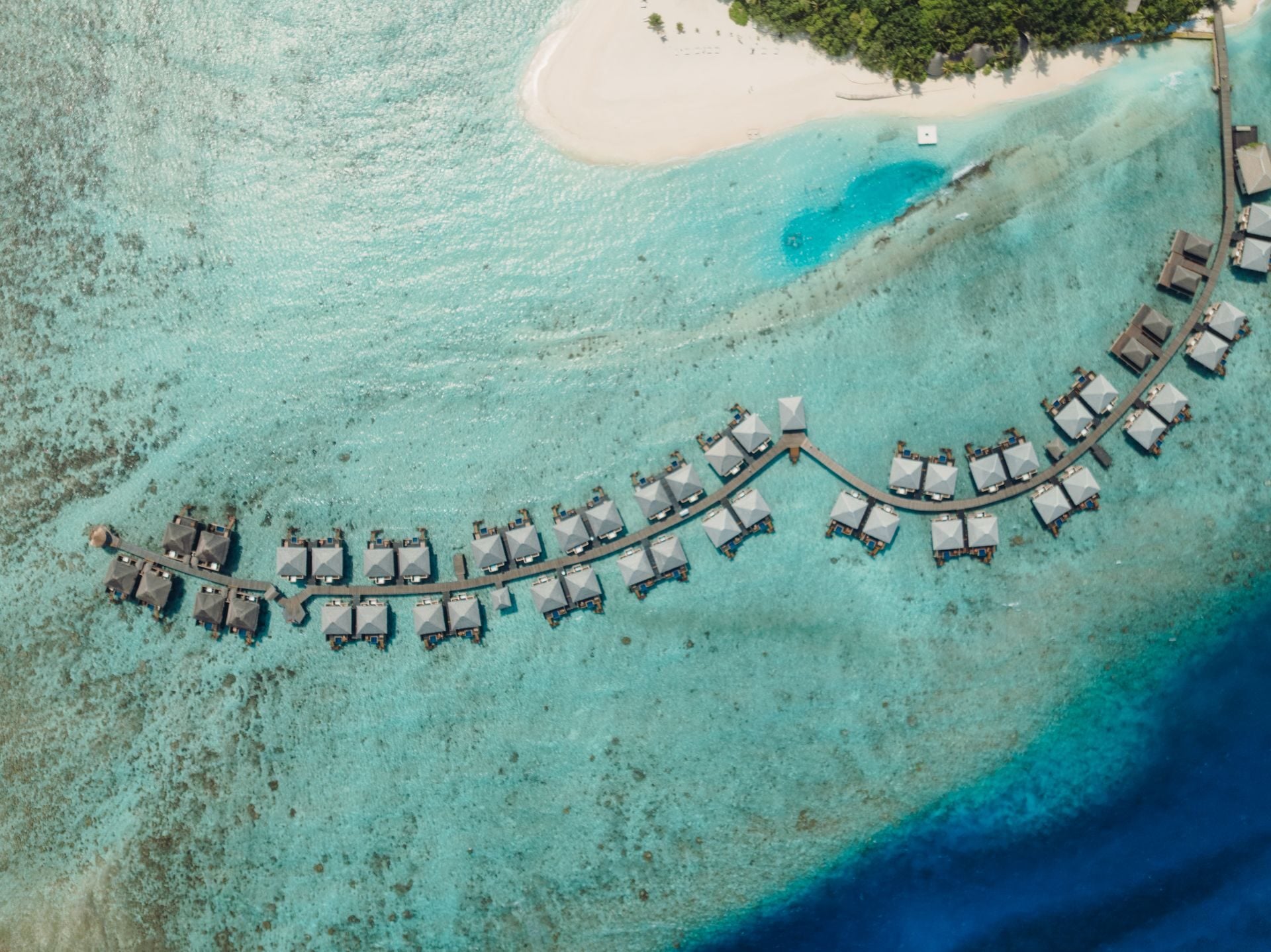 Luftaufnahme von oben auf Wasservillen mit Plunge-Pools entlang eines geschwungenen Holzstegs.