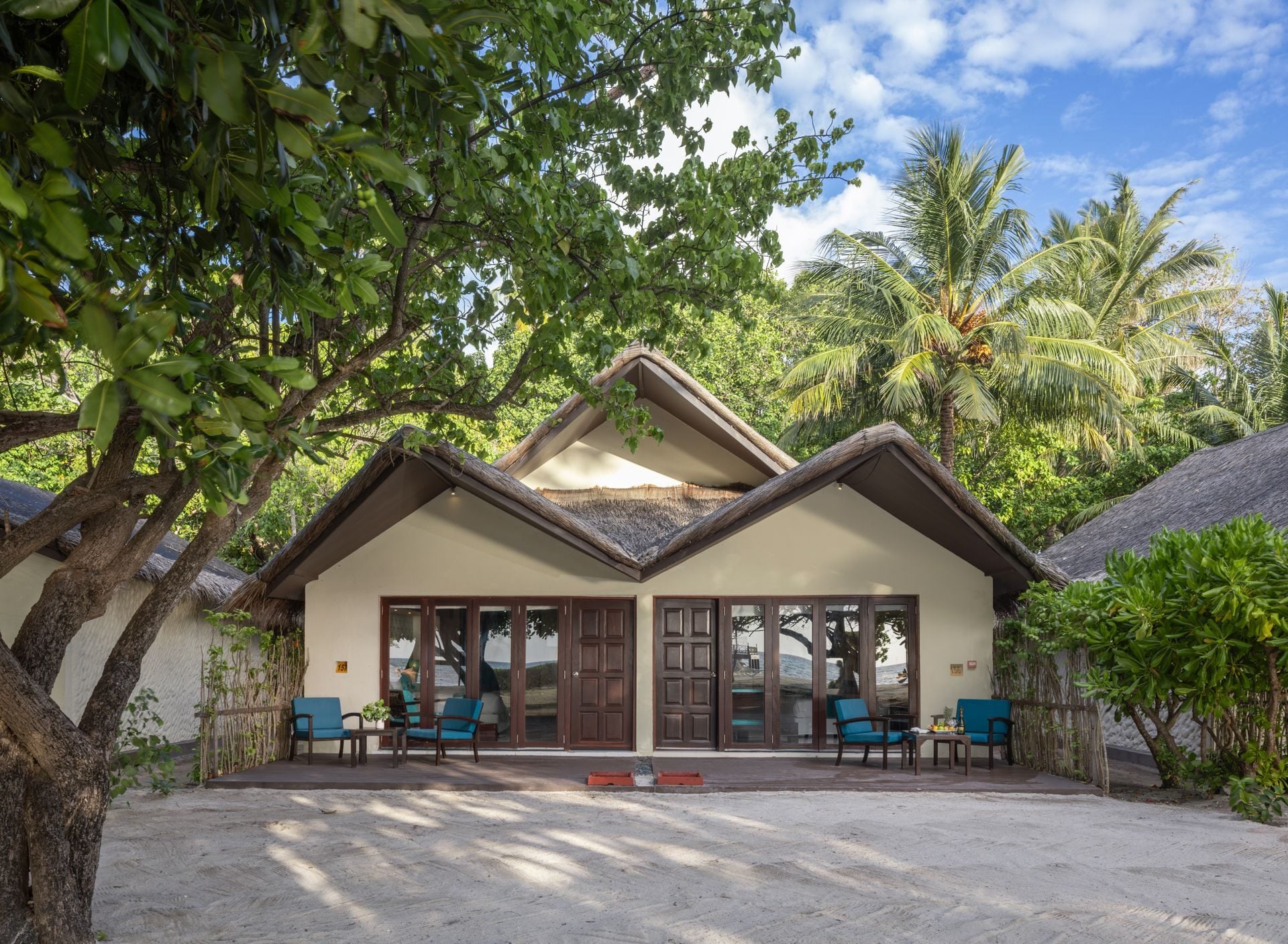 Außenansicht einer Villa mit Reetdach, schattiger Terrasse und blauen Sesseln, umgeben von Bäumen.
