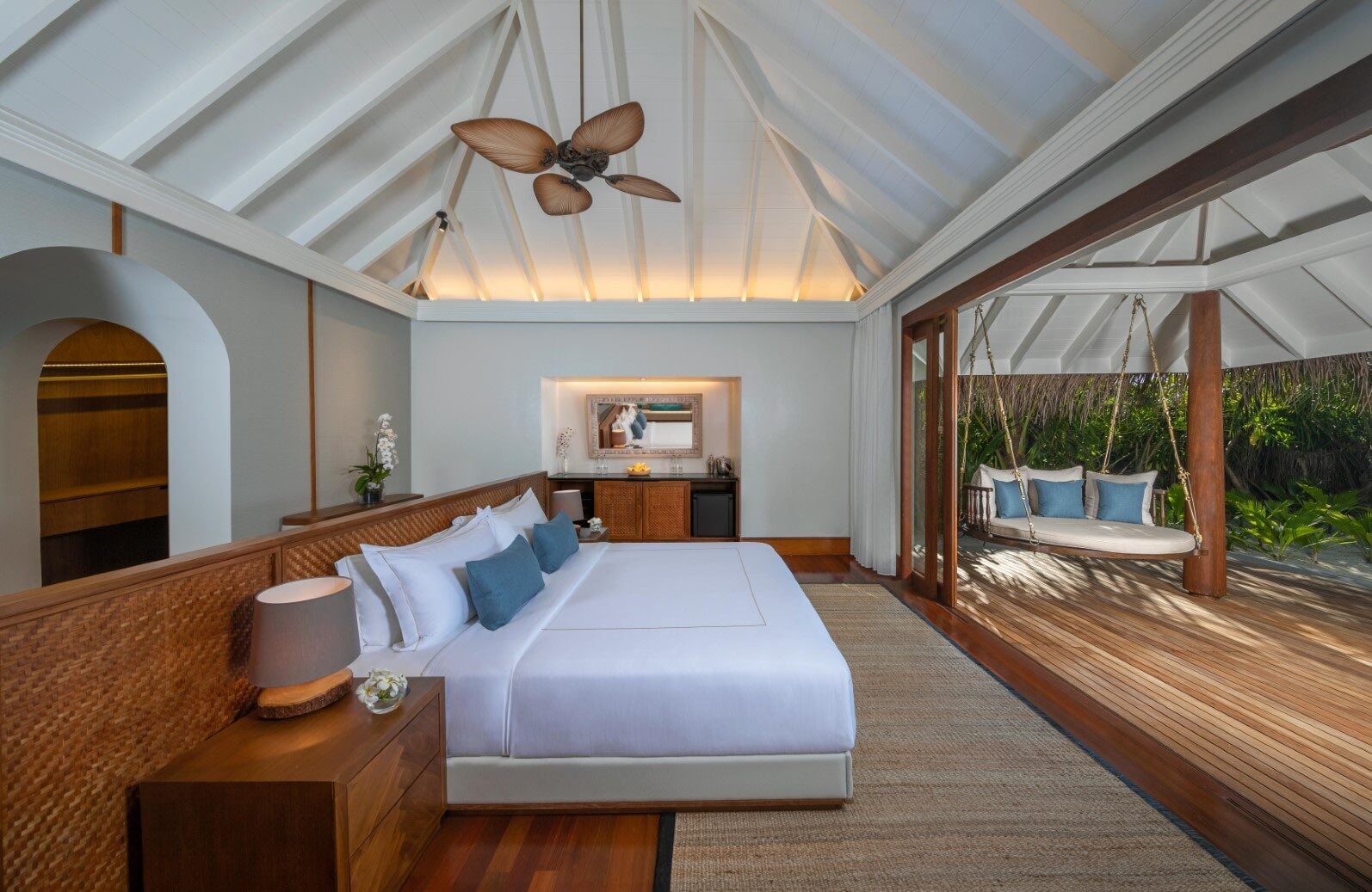 Ein geräumiges Villa-Schlafzimmer mit weißem Bett, Deckenventilator und offenen Türen zu einer Holzterrasse mit Hängebett.