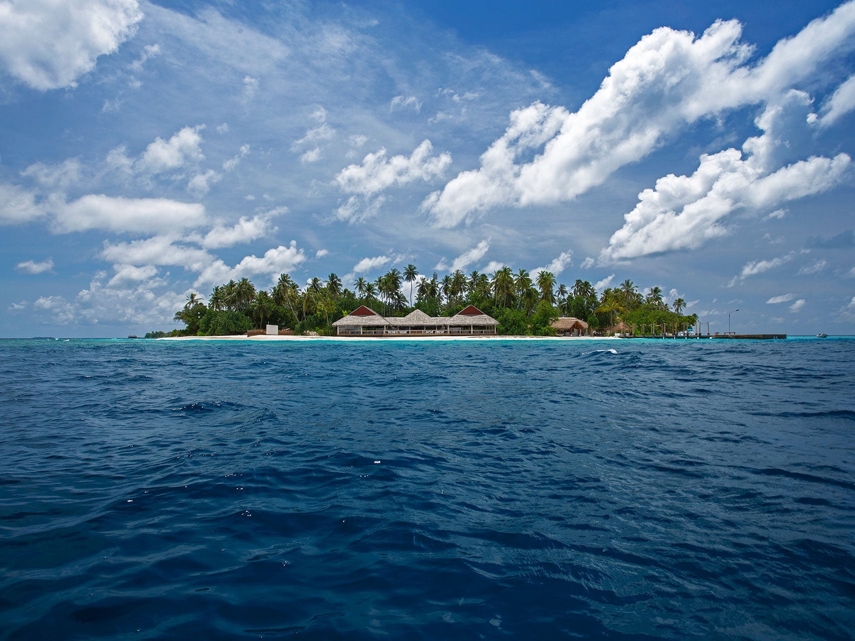 Blick übers Meer auf eine kleine, palmenbewachsene Insel mit niedrigen Gebäuden am Wasser.