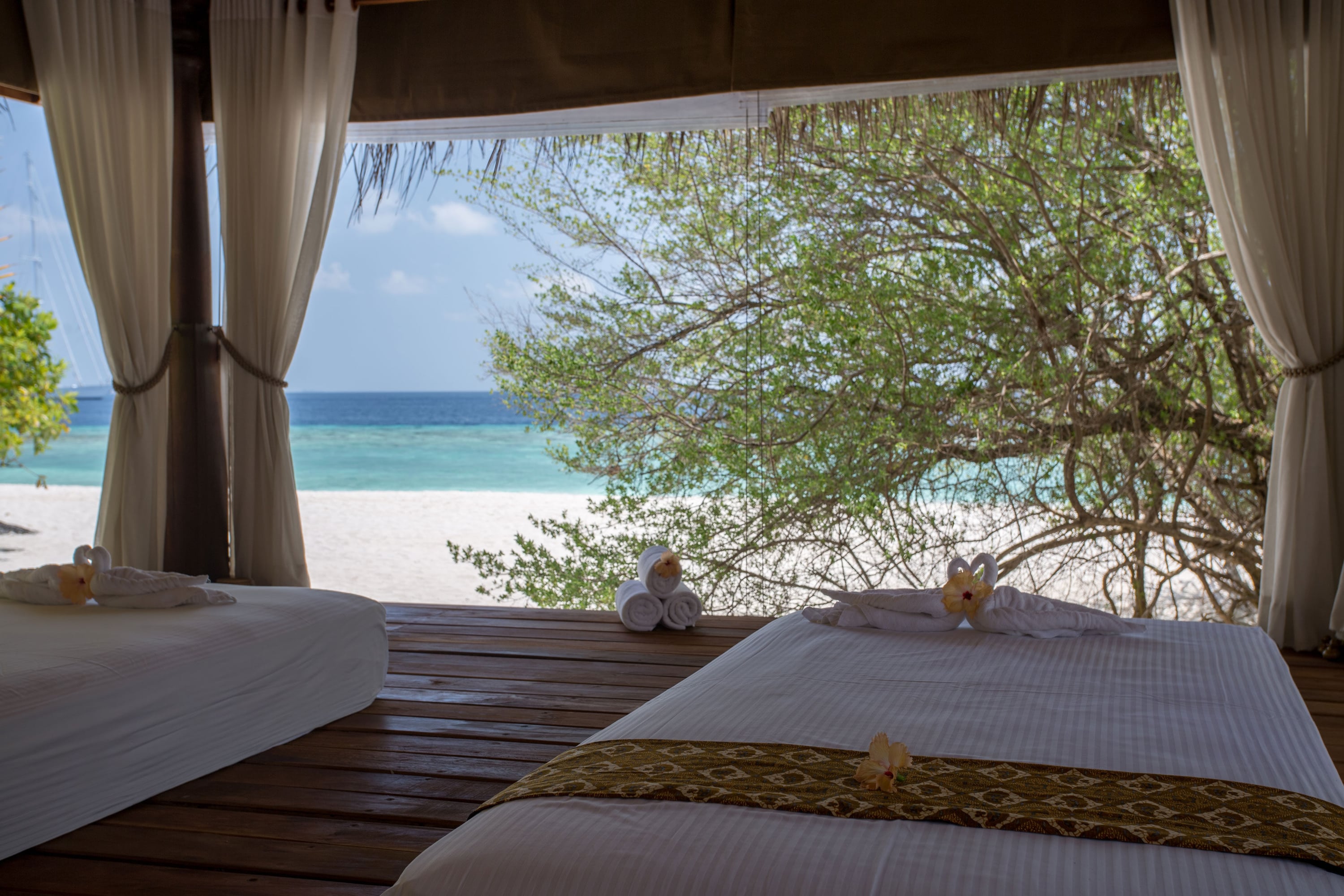 Zwei Massageliegen auf einer Holzterrasse mit offenem Blick auf Strand und türkisfarbenes Meer.