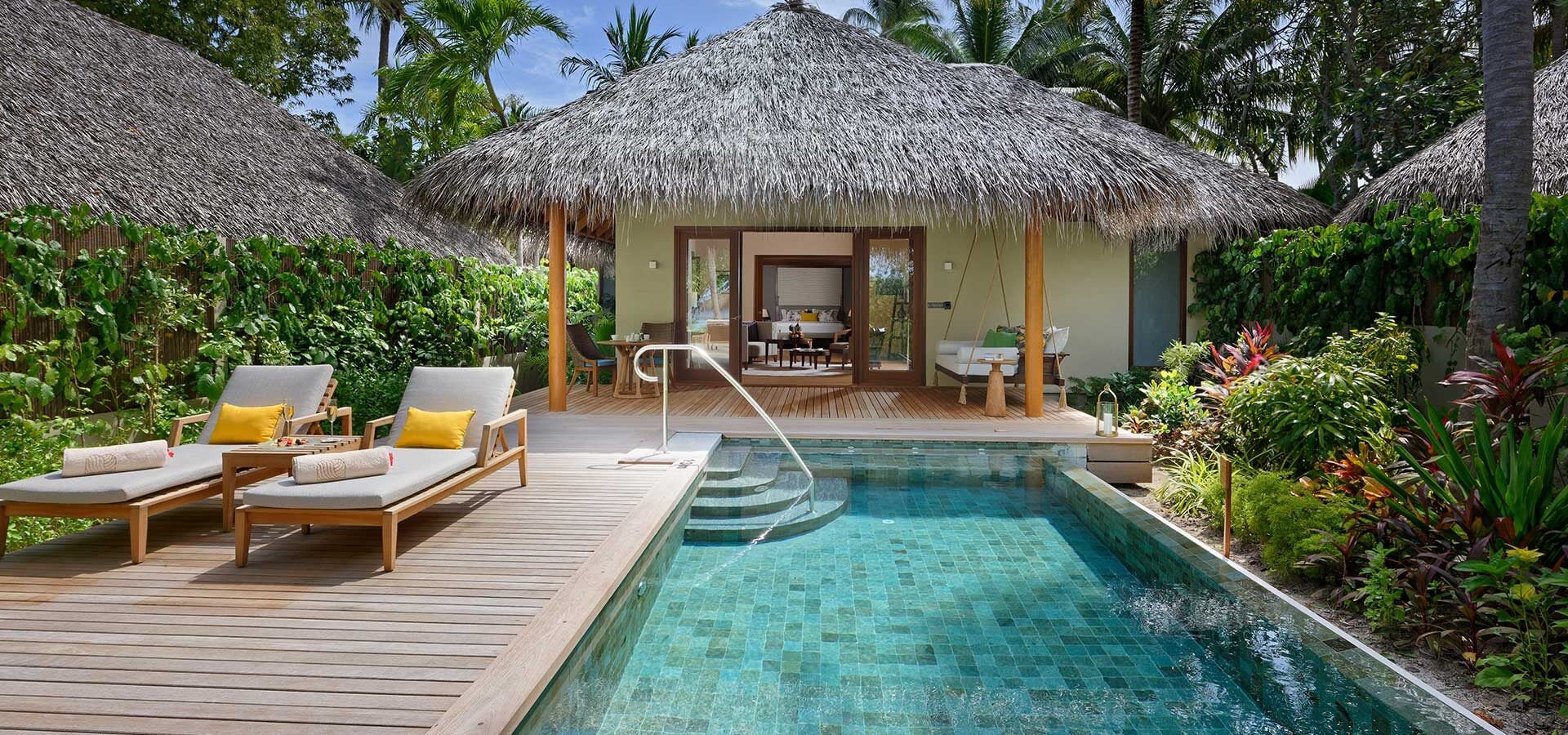 Ein privater Plunge Pool mit Liegen befindet sich auf einer Holzterrasse vor einer strohgedeckten Villa, umgeben von tropischem Grün bei Baros Maldives.
