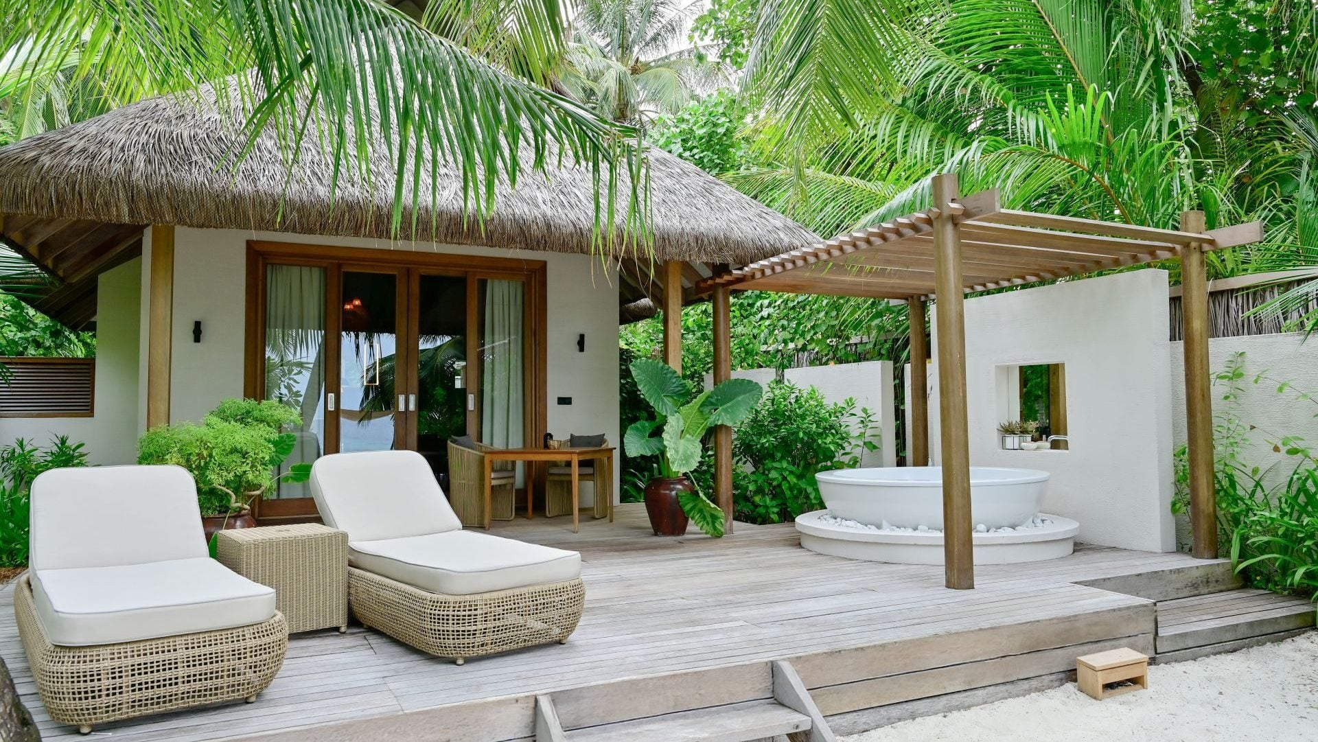 Die Terrasse einer Strandvilla bietet Liegen, einen Esstisch und eine runde Außenbadewanne, umgeben von tropischem Grün.