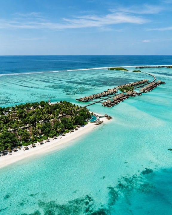 Aerial view of a tropical island in the Maldives, featuring lush greenery and a white sandy beach. Wooden overwater bungalows extend into the clear turquoise ocean. The vibrant reef and deep blue sea 
