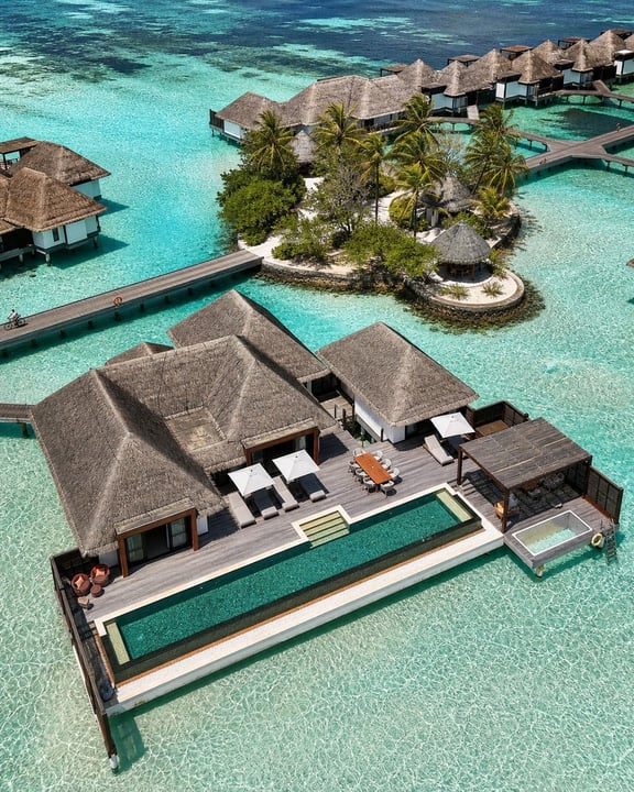 Luftaufnahme luxuriöser Overwater-Bungalows und privater Villen, verbunden durch hölzerne Stege, umgeben von klarem türkisfarbenem Wasser. Im Hintergrund eine kleine Insel mit Palmen.