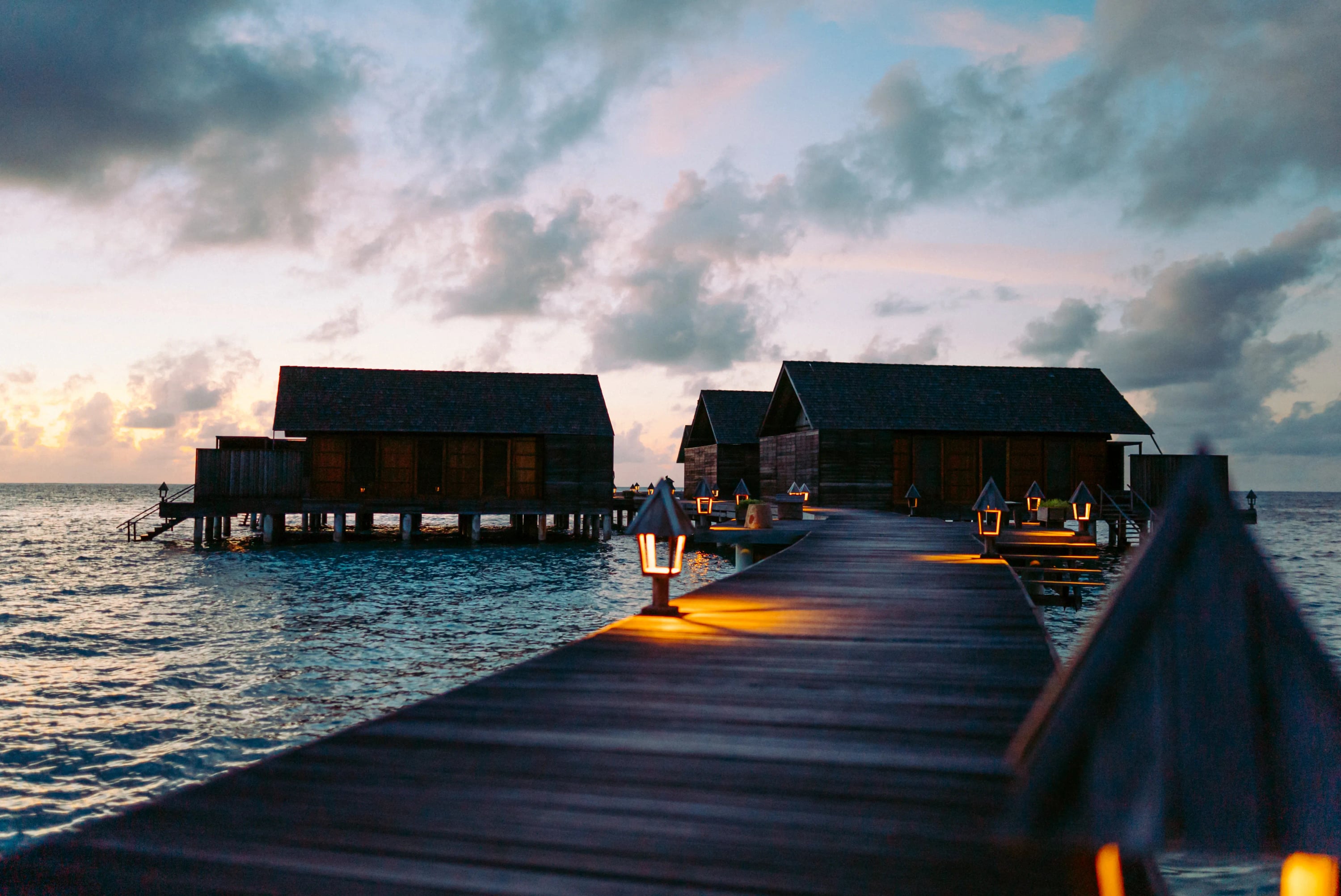 La jetée menant à l'Overwater Villa Deluxe au crépuscule, lampes allumées le long du ponton à Gangehi Island Resort & Spa