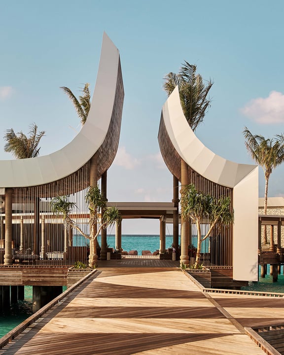 L'emblématique pavillon d'arrivée de .Here Baa Atoll, aux Maldives, avec ses formes architecturales courbes et élancées, une passerelle en bois au-dessus d'une eau cristalline, des palmiers tropicaux et une vue panoramique sur l'océan Indien en