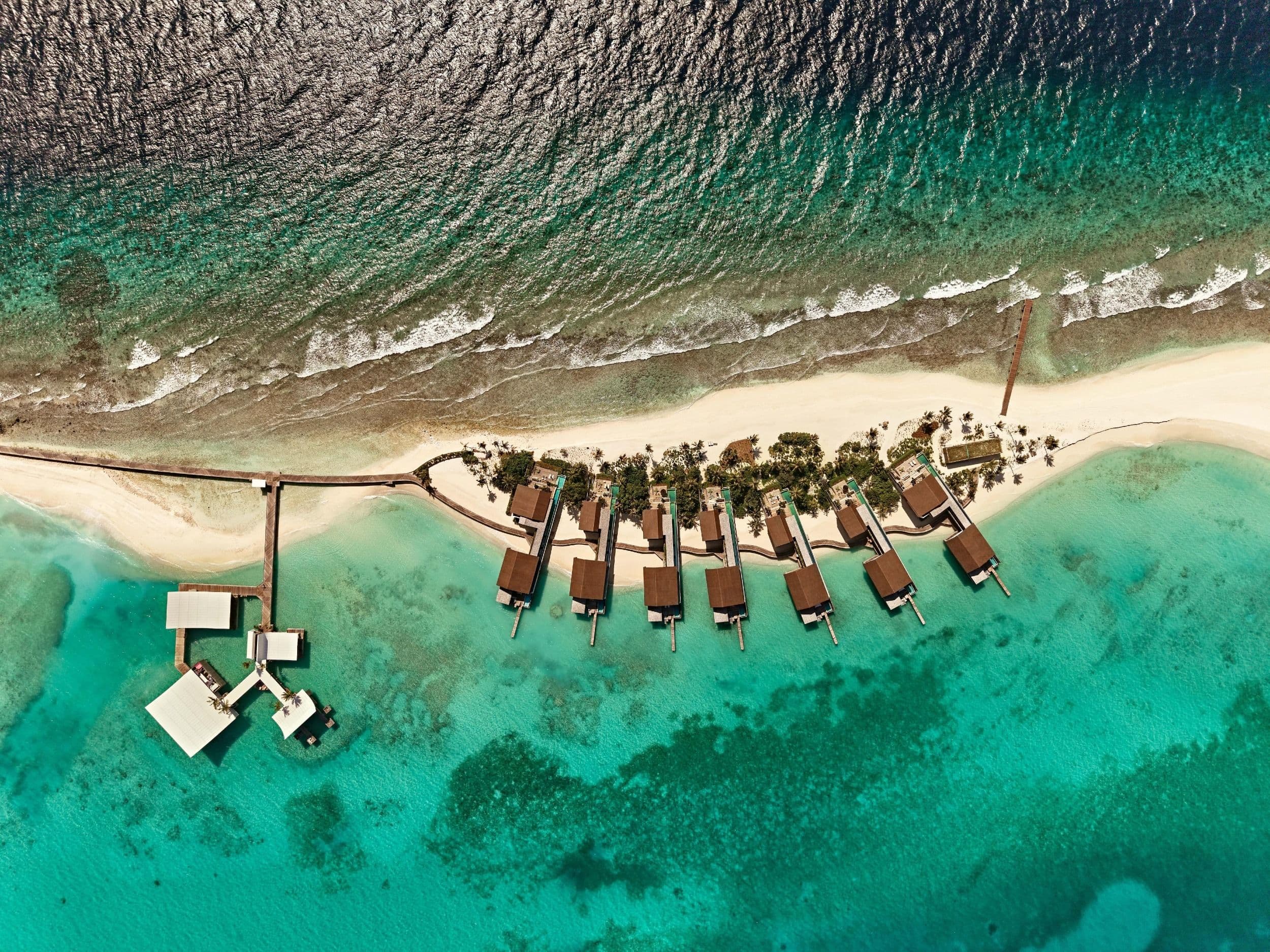 Une vue aérienne d'une île étroite bordée de Residences le long de sa plage, avec un lagon d'un côté et l'océan ouvert de l'autre.