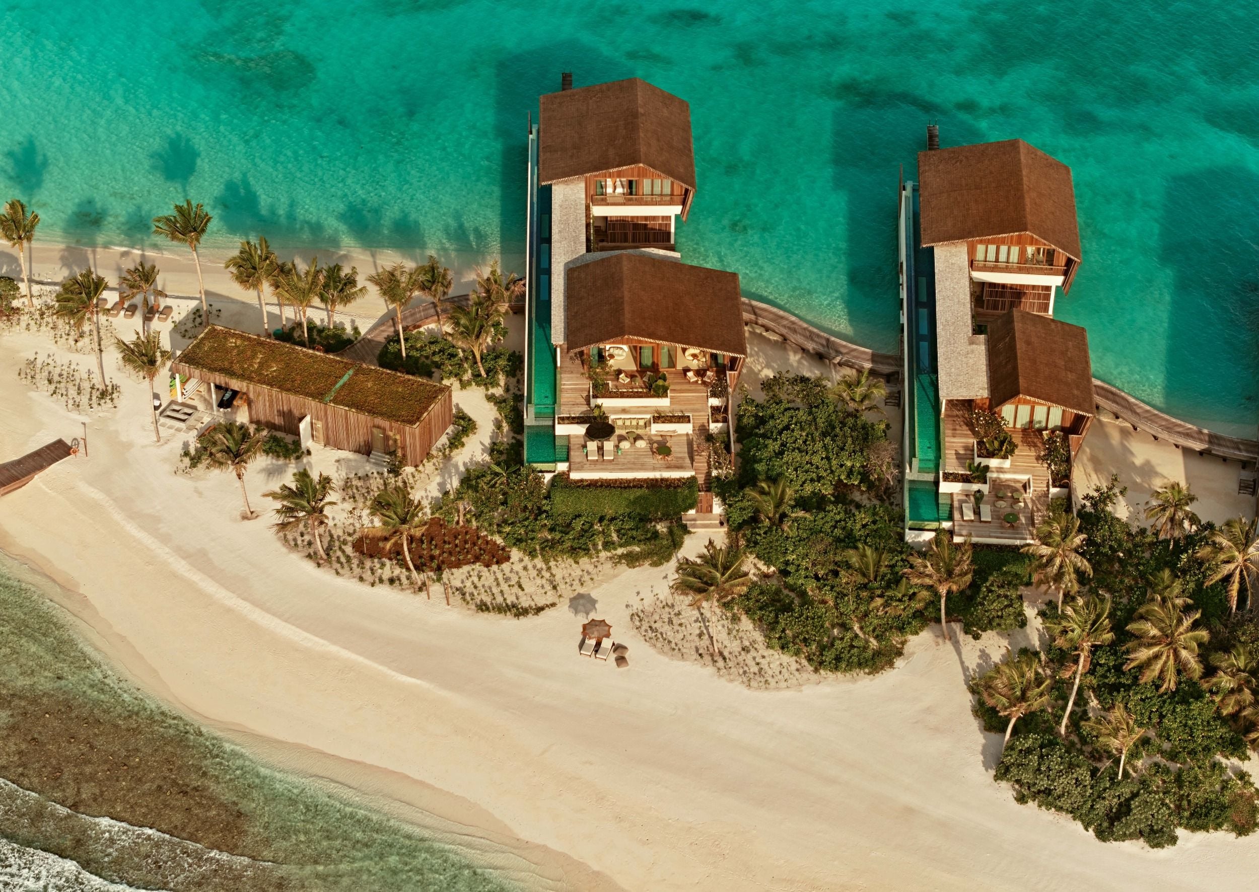 Une vue plongeante sur deux Residences à plusieurs étages dotées de longues piscines vitrées, nichées parmi les palmiers au-dessus d'une plage de sable blanc.