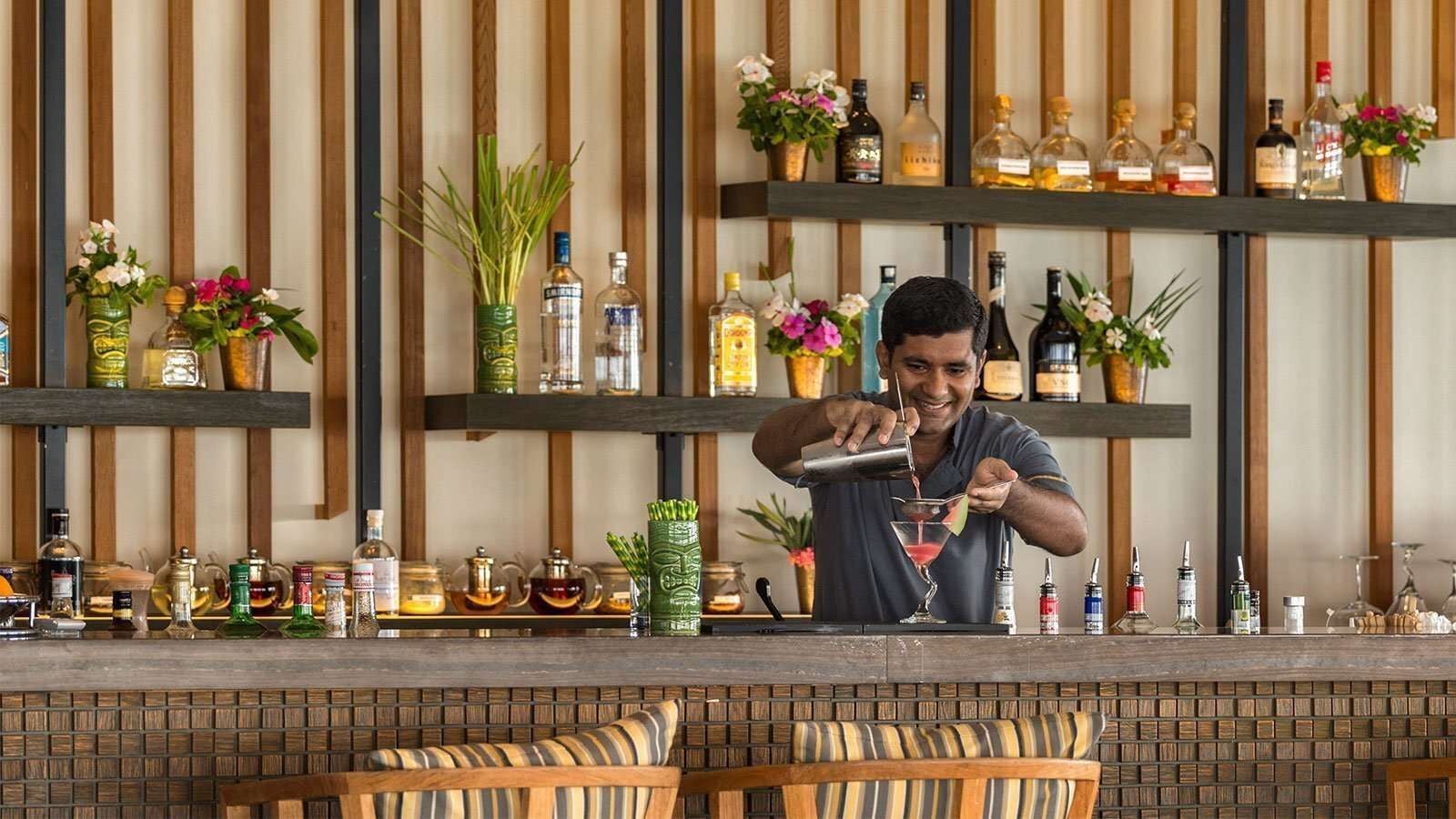 Ein Barkeeper schenkt an einer mit Flaschen und Blumen bestückten Holzbar im Hurawalhi Island Resort einen Cocktail aus dem Shaker ein.