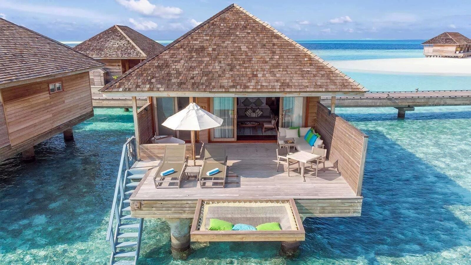 Eine Terrasse einer Wasservilla mit Sonnenliegen, Sonnenschirm und einer Netzhängematte über klarem türkisfarbenem Lagunenwasser.