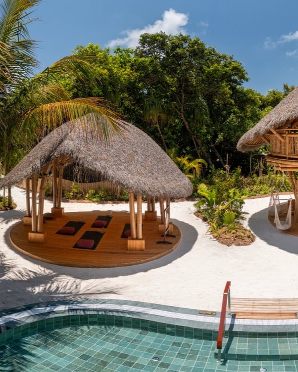 Un cadre tropical en plein air avec un pavillon de yoga au toit de chaume et des tapis sur une terrasse en bois, entouré d’une végétation luxuriante. À proximité, une petite hutte ronde surélevée abrite un hamac. Au premier plan, le