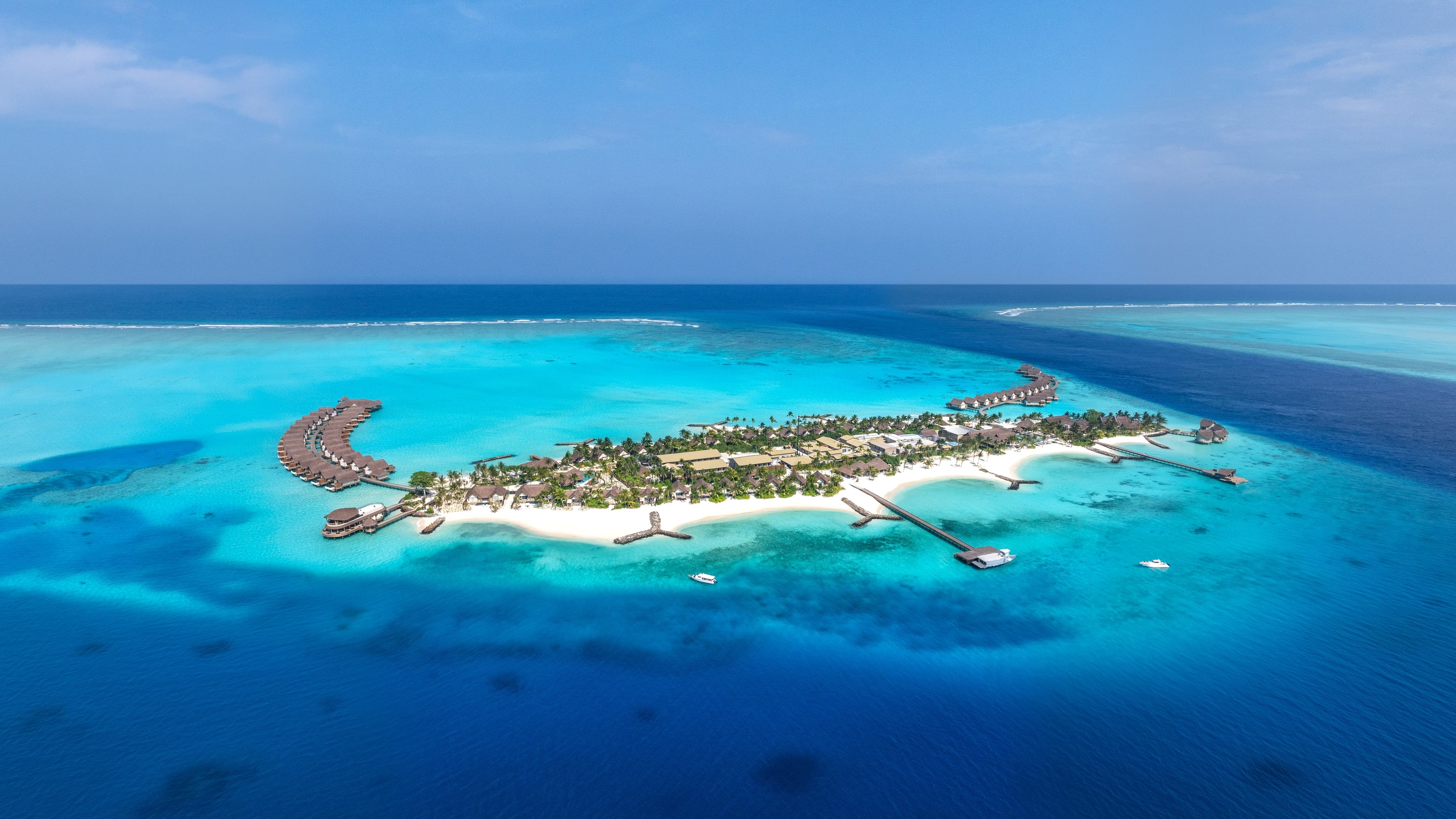 Aerial of the whole island, with an arc of overwater villas at each end and the arrival jetty reaching into the lagoon.