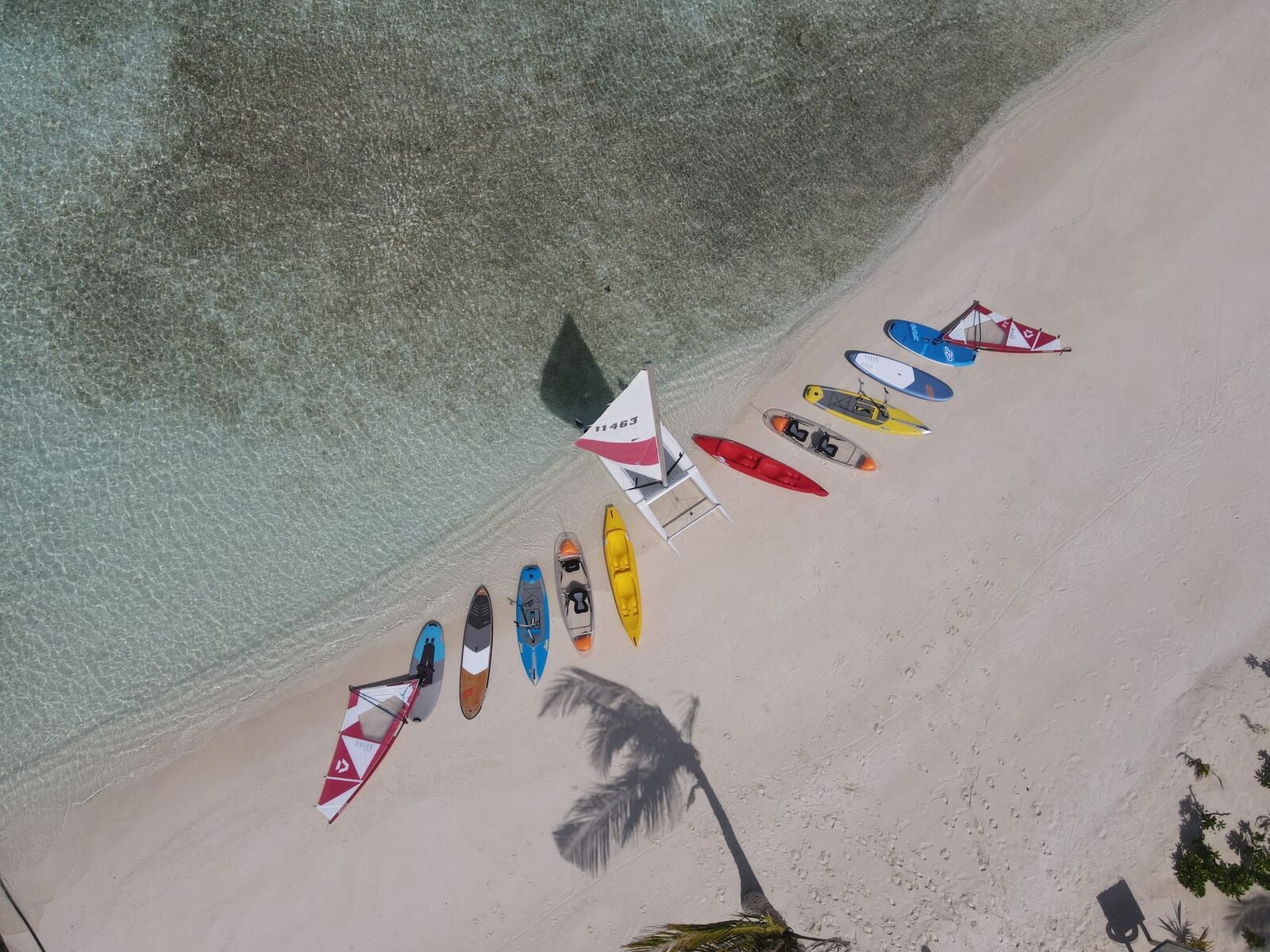 Luftaufnahme von Kajaks, Paddleboards und Segelbrettern, aufgereiht an einem weißen Sandstrand neben türkisfarbenem Wasser.