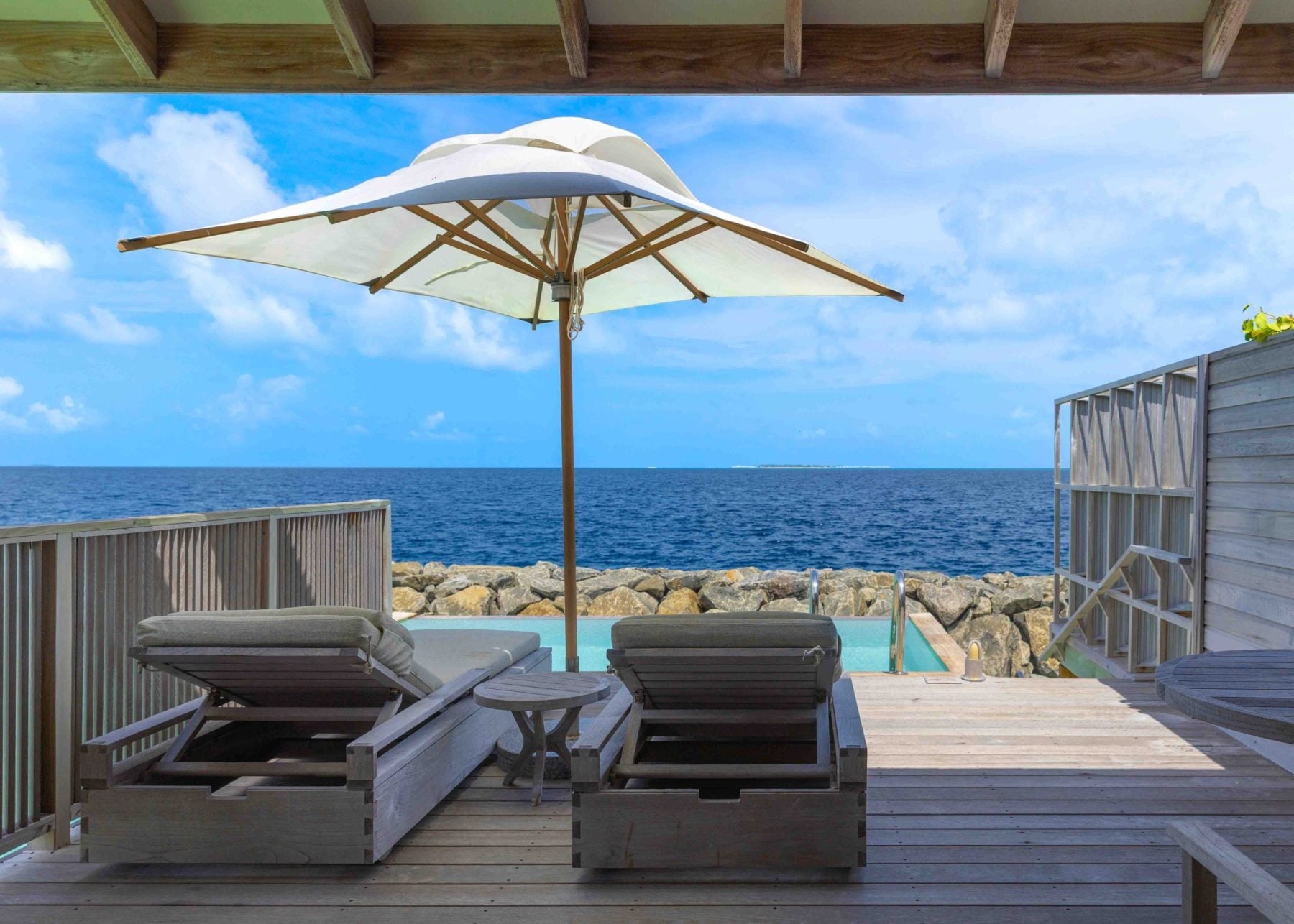 Zwei Sonnenliegen und ein Sonnenschirm auf einer hölzernen Terrasse über dem Wasser mit einem kleinen Plunge-Pool und Meerblick.