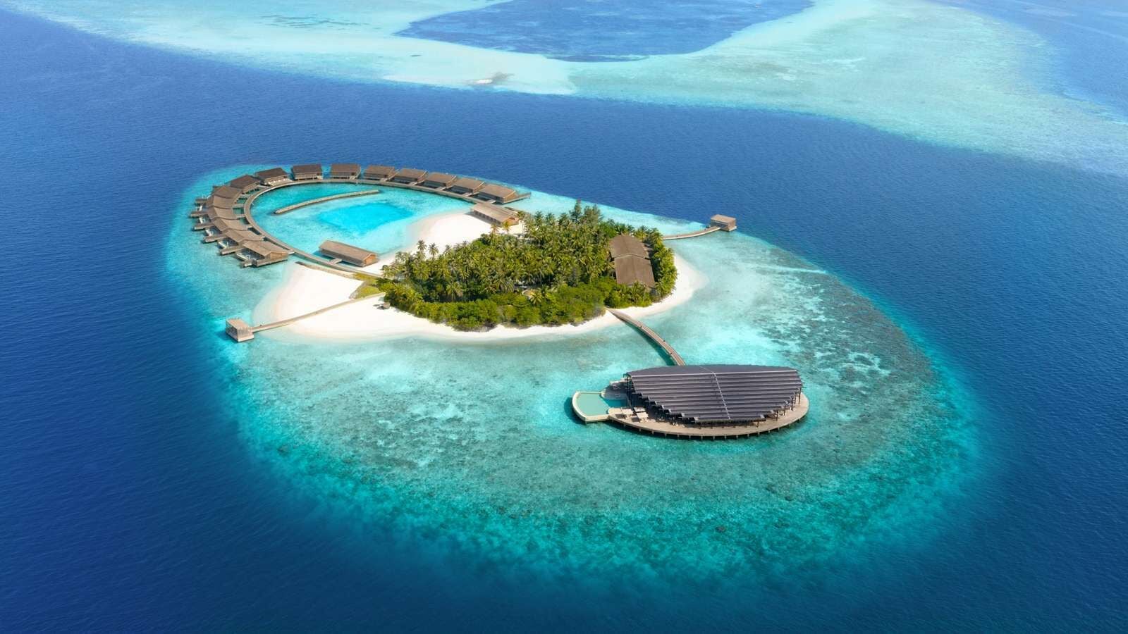 Luftaufnahme einer ringförmigen Resortinsel mit Wasservillen, einem zentralen Lagunenpool und umgebendem Korallenriff.