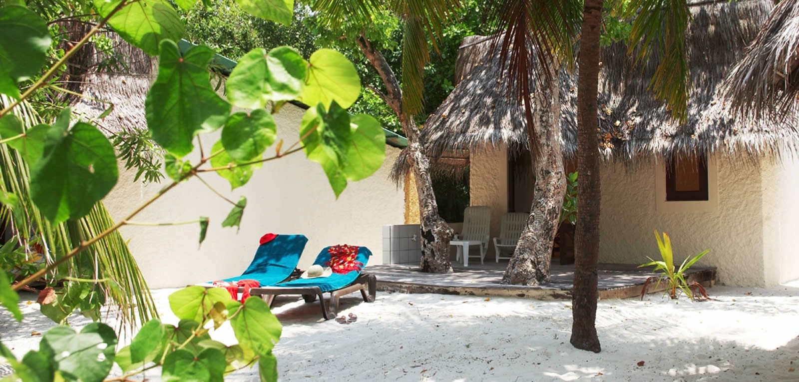 Sonnenliegen mit Handtüchern stehen vor einem strohgedeckten Beach Bungalow im Schatten von Palmen.