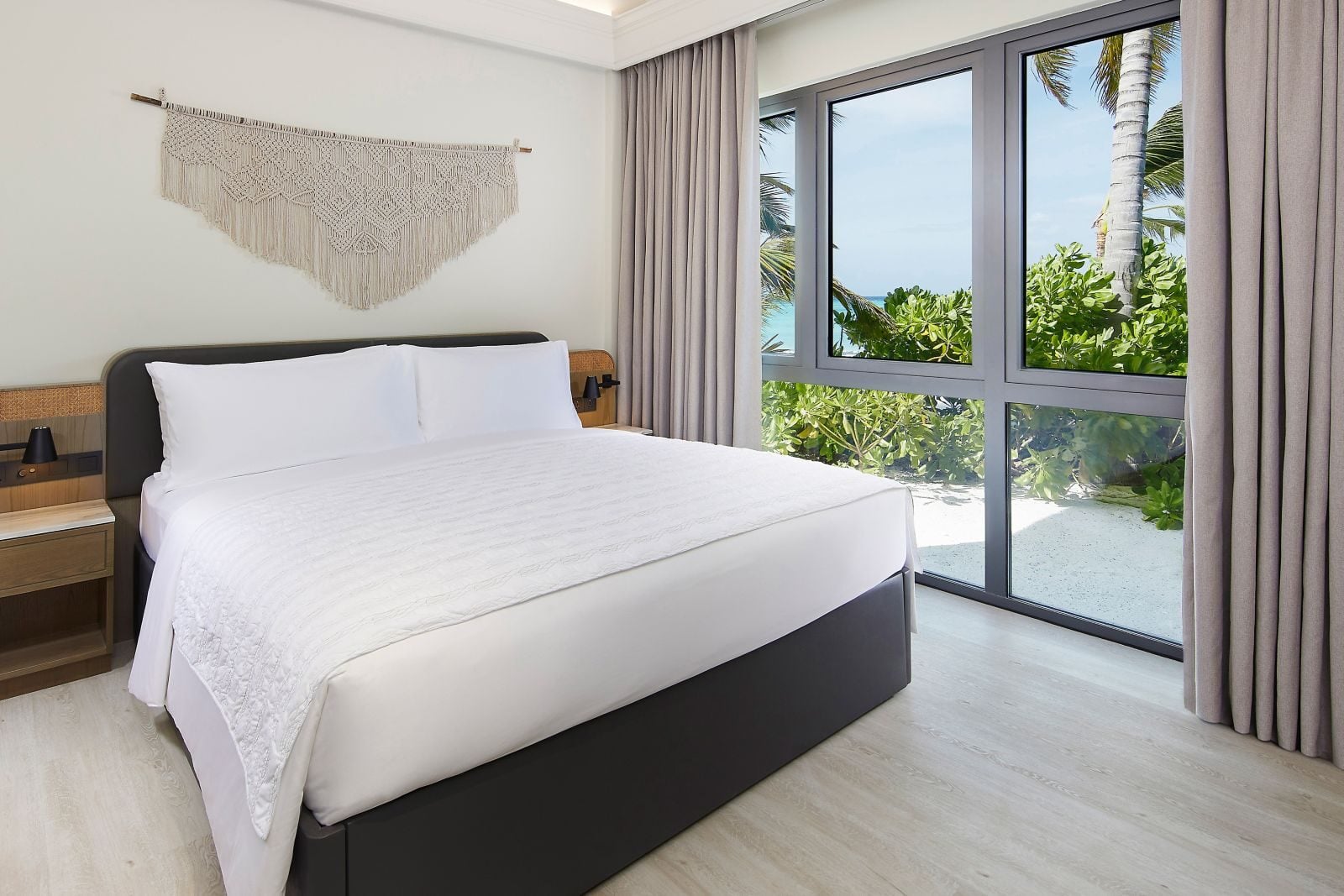 Ein helles Schlafzimmer mit großem weißen Bett liegt Glastüren mit Blick auf Strand und Palmen gegenüber.