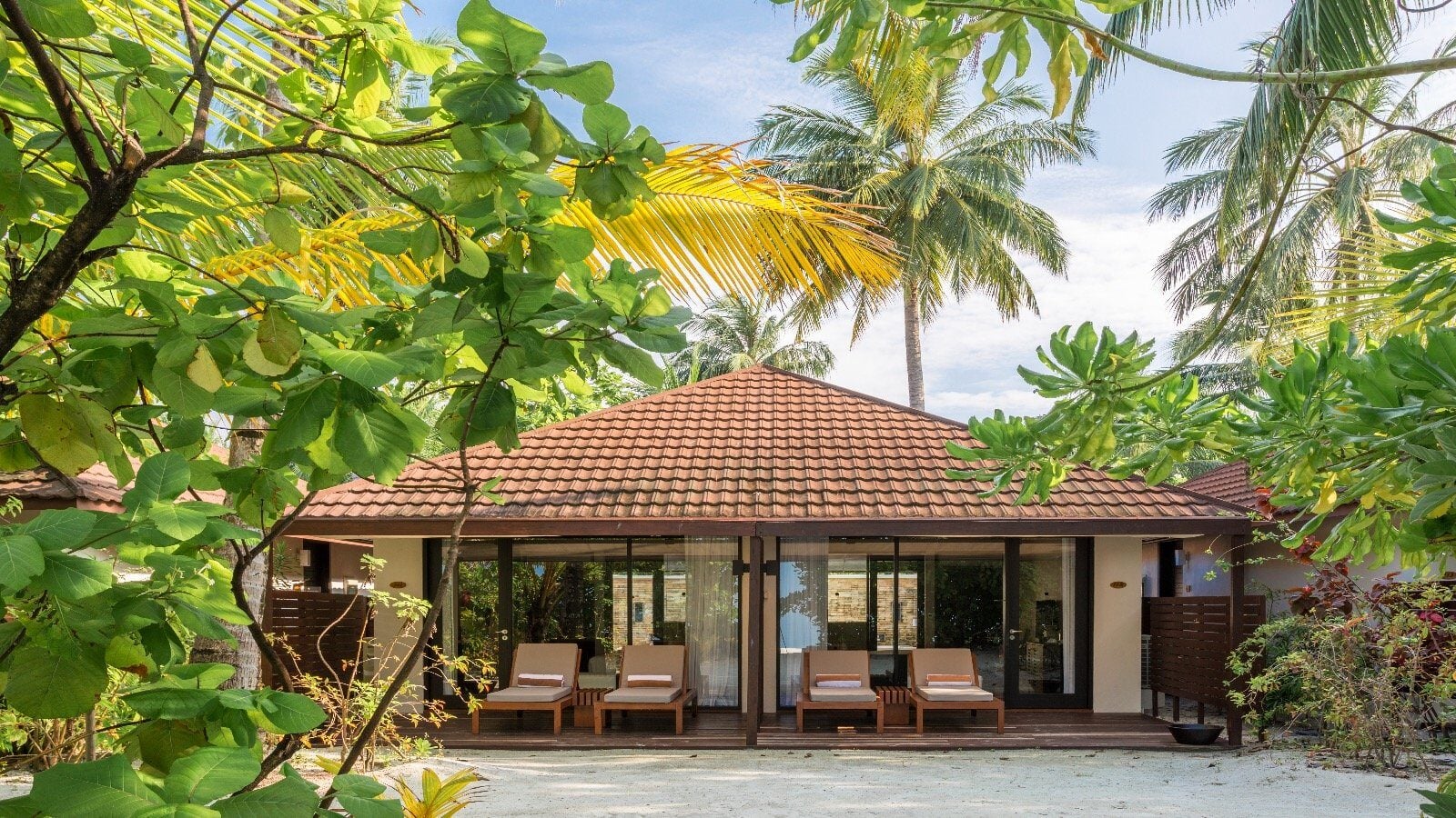 Eine Strandvilla mit Ziegeldach und Liegen auf der Terrasse, umgeben von tropischem Grün.