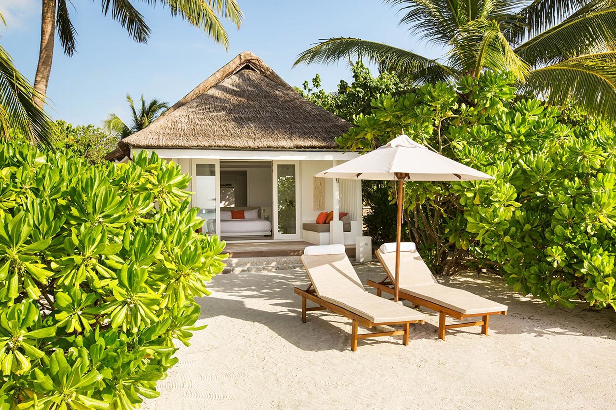 Ein strohgedecktes Beach Pavilion liegt inmitten üppigen Grüns, davor zwei Sonnenliegen und ein Sonnenschirm im Sand.