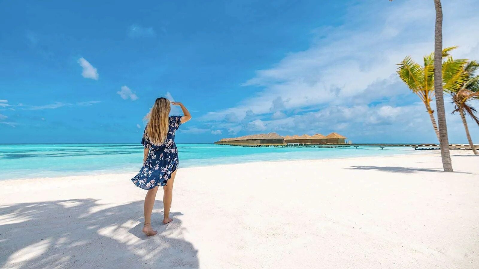 Eine Frau in einem geblümten Kleid steht am weißen Sandstrand und blickt über die Lagune auf ein überwasserbauwerk.