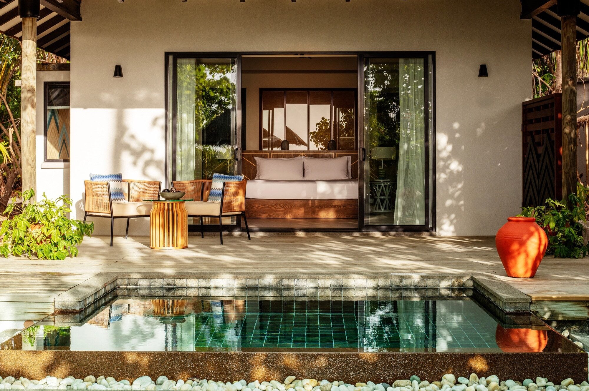 Ein privater Plunge Pool auf einer Holzterrasse vor dem Schlafzimmer einer Villa mit offener Schiebetür und Sitzbereich.