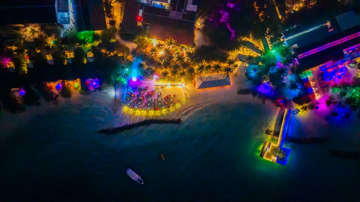 Aerial nighttime view of Oaga Art Resort in the Maldives, showing brightly lit pathways, villas, and communal spaces glowing in multiple colors. The island appears active and vibrant, with illuminated