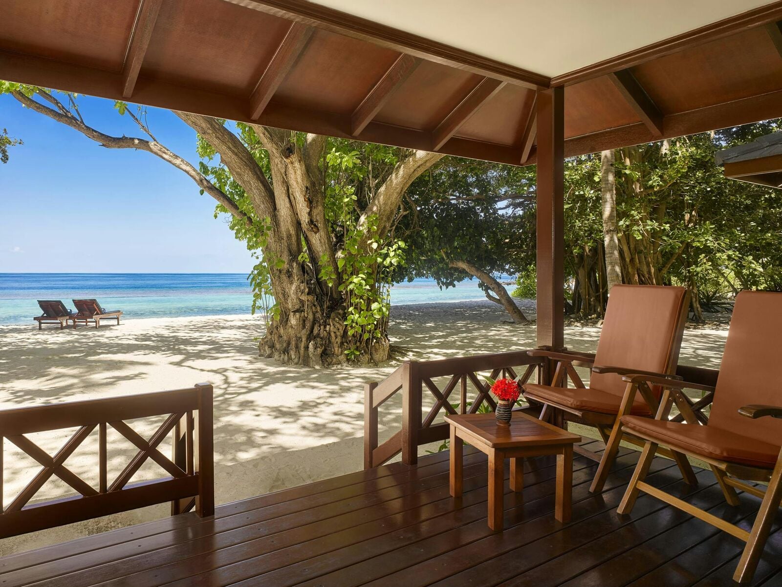 Eine schattige Holzterrasse mit Liegen und einem Tisch bietet durch Bäume hindurch Blick auf einen Sandstrand und türkisfarbenes Meer.