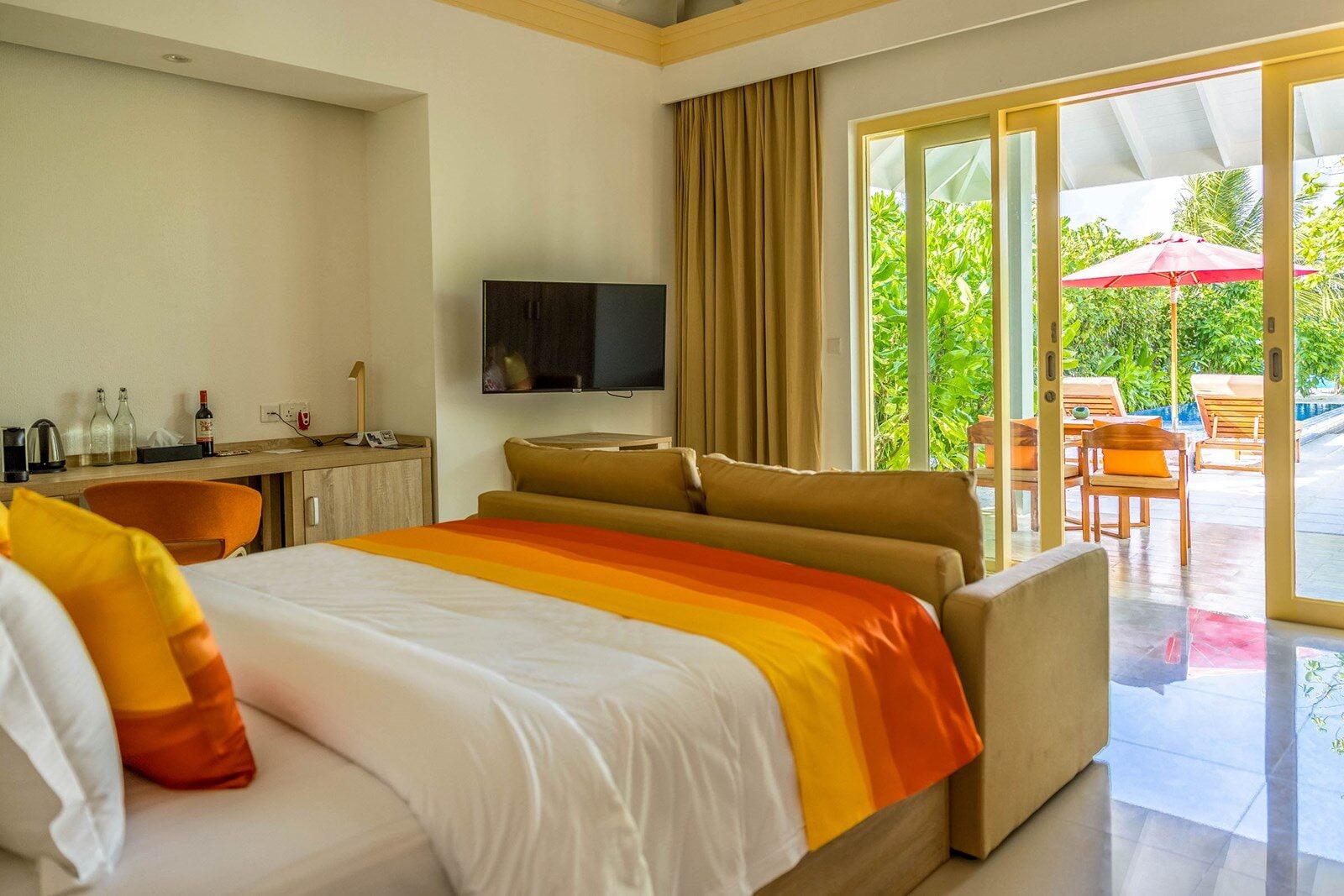 Ein Hotelschlafzimmer mit orange gestreifter Tagesdecke öffnet sich zu einer Terrasse mit Sofas und Poolblick.