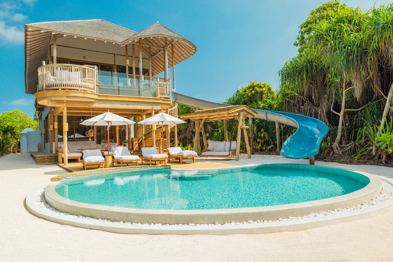 Eine zweistöckige Strandvilla mit geschwungenem Pool, Sonnenliegen und einer blauen Rutsche, die ins Wasser führt.