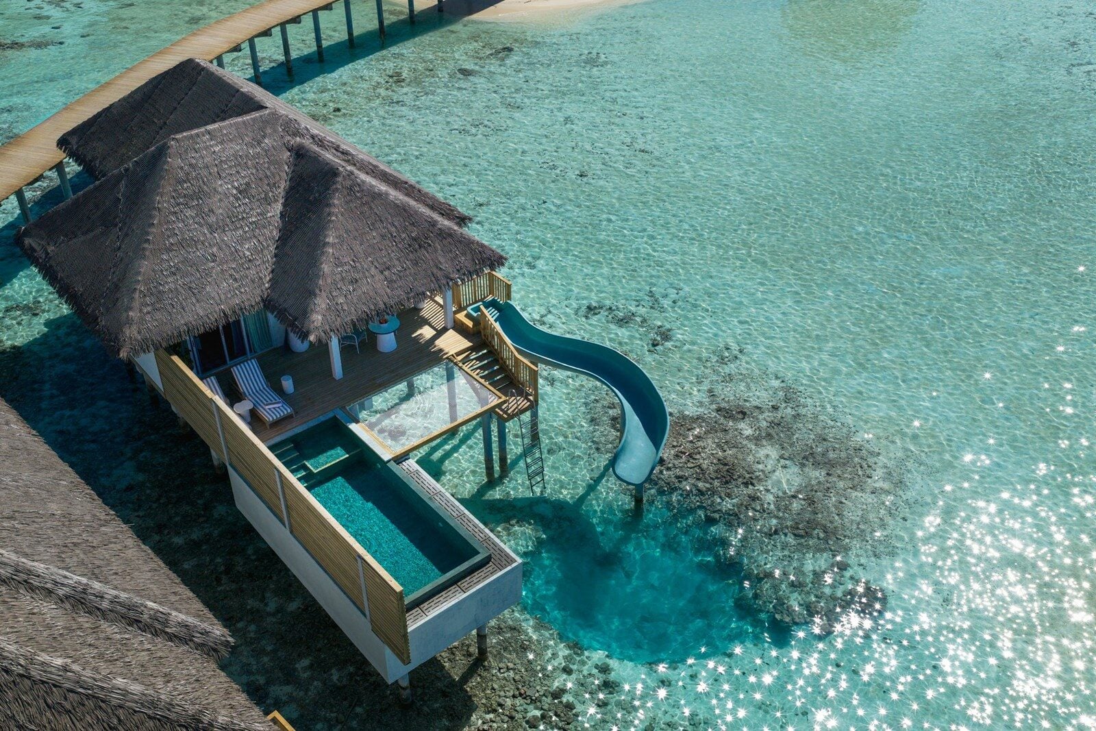 Luftaufnahme einer strohgedeckten Wasservilla mit privatem Plunge-Pool und einer Rutsche, die in die Lagune führt.