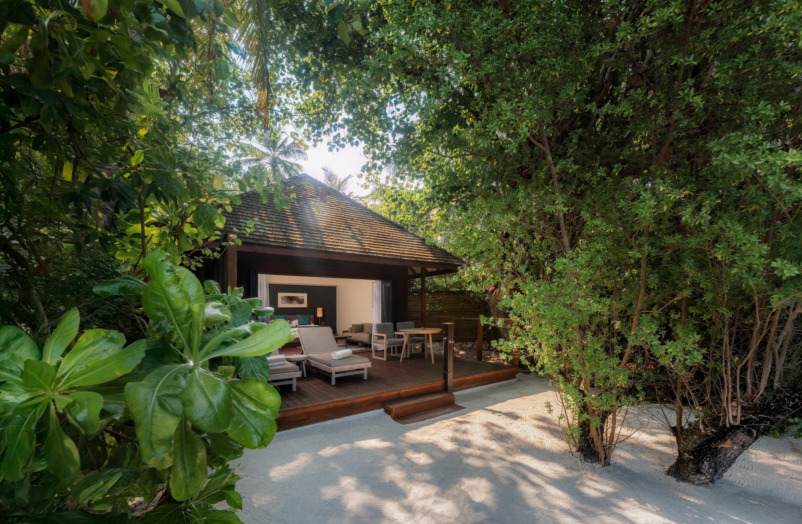 Eine Villa mit Reetdach, einer Holzterrasse und Liegestühlen ist von dichtem tropischem Grün umgeben.