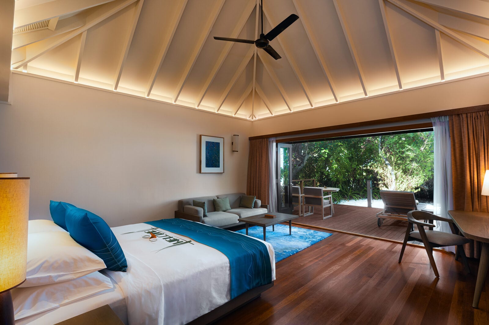 Ein helles Villa-Schlafzimmer mit Kingsize-Bett, Sitzbereich und einer Holzterrasse, die sich zum Grün nach draußen öffnet.