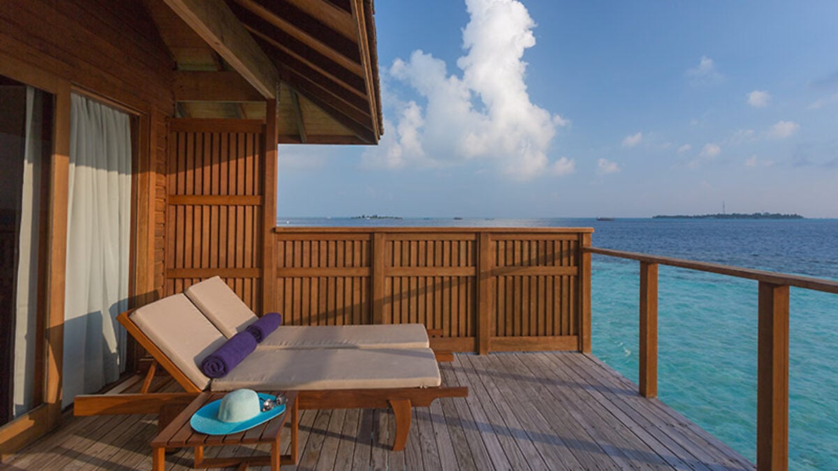 Ein hölzernes Sonnendeck einer Wasservilla mit zwei Liegestühlen und einem Hut, mit Blick auf die türkisfarbene Lagune im Vilamendhoo Island Resort & Spa.