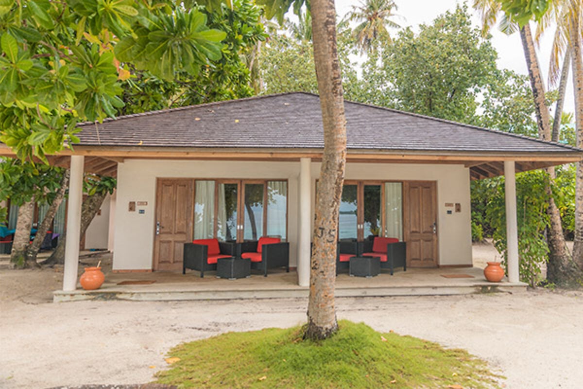 Eine Strandvilla mit spitzem Reetdach und schattiger Terrasse liegt zwischen Palmen auf sandigem Grund.