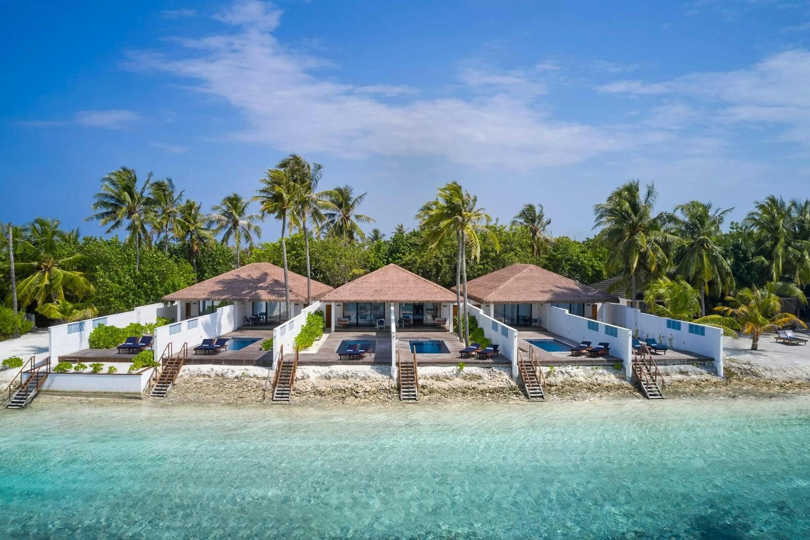 Drei Beachfront Villas mit privaten Tauchbecken stehen in einer Reihe entlang des Sandstrands, umgeben von Palmen.